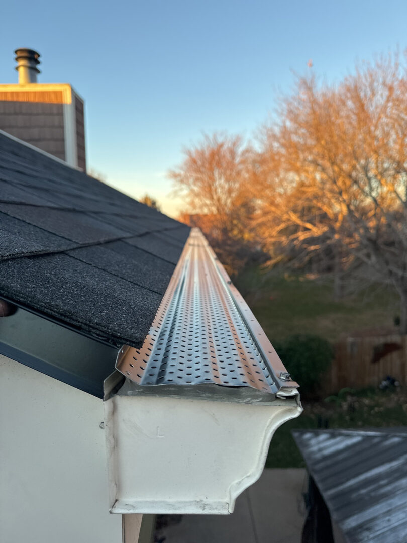 The photo shows a close-up view of a house's exterior, focusing on a white gutter with a leaf guard installed on top. The leaf guard has a diamond-shaped mesh pattern designed to allow water through while preventing debris from entering the gutter. The roof has dark shingles, and in the background, there's a chimney and a tree with golden leaves, indicating it might be autumn. The image seems to be taken from inside through a window, as reflected elements can be seen on the glass surface. This photo is likely showcasing the gutter installation work by Pillar Exteriors, a company specializing in home exterior services.