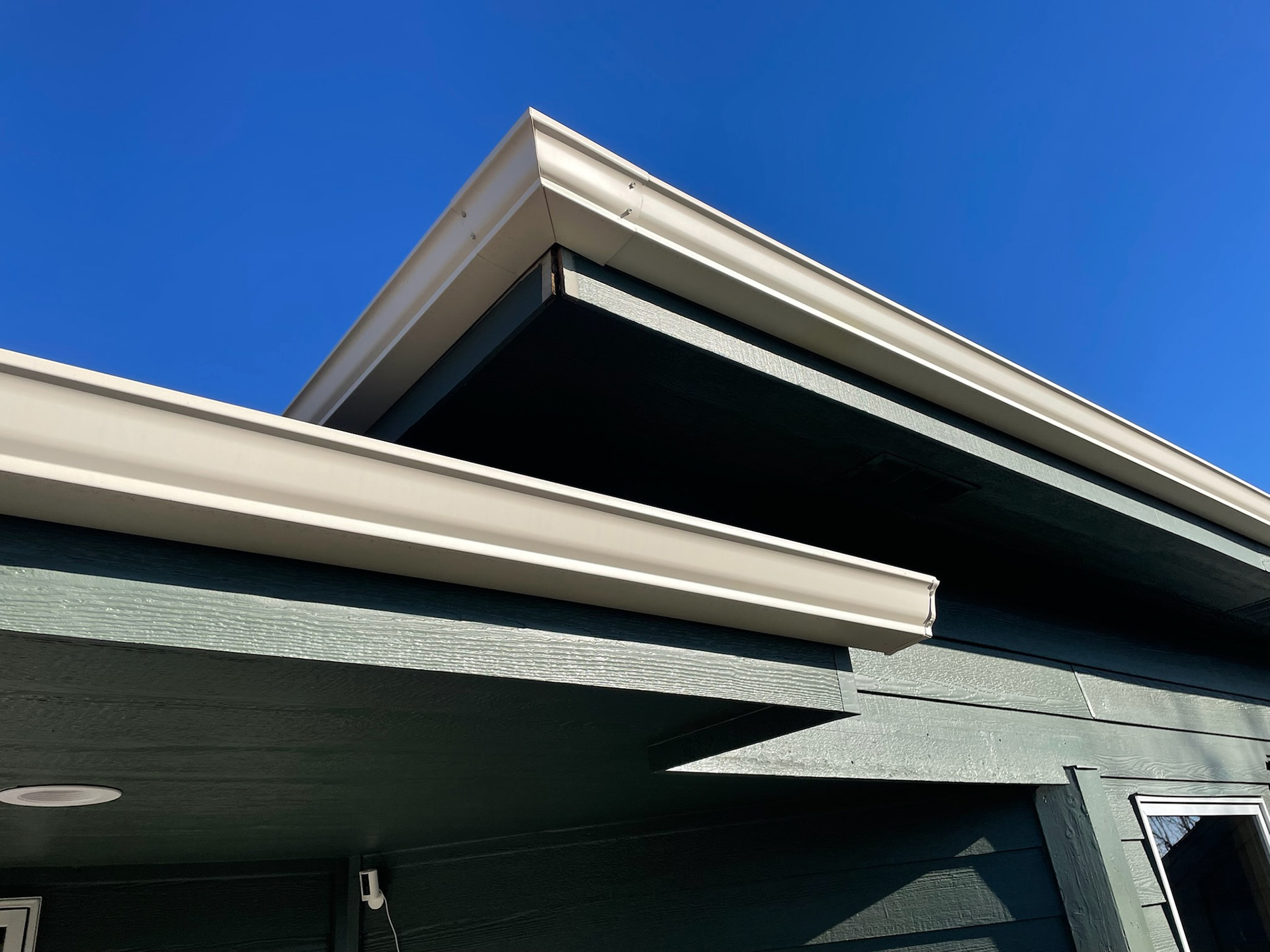 The image shows a close-up view of a building's roofline against a clear blue sky. The roof features beige soffit panels and matching fascia. There's a sharp geometric interplay between the different angles of the roof edges and overhangs. The structure appears modern with clean lines and a minimalist aesthetic. This photo seems to be taken from a low angle, looking up towards the sky, and highlights the crisp and professional finish of the roofing work likely completed by Pillar Exteriors.
