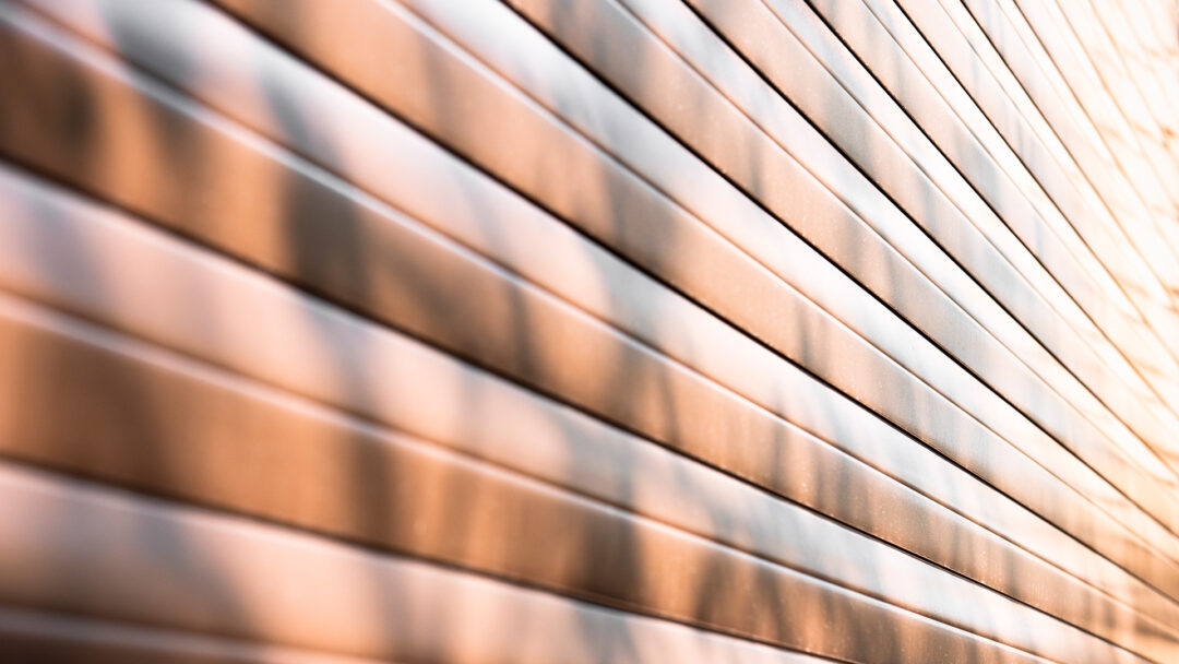 The image is a close-up of metal siding with a horizontal orientation, showcasing a series of clean, straight lines that create a pattern of alternating light and shadow. The metallic finish of the siding panels reflects light, giving the surface a luminous quality with warm tones. The perspective and focus on the textural details highlight the material's durability and sleek appearance.

The image could be used by Pillar Exteriors to exemplify their offerings in metal siding options. It conveys a sense of modernity and strength, qualities often sought after in siding materials for contemporary building exteriors. The play of light across the panels also suggests the aesthetic appeal of such materials, which can add a visually striking element to a building's design.